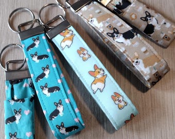 May include: Several keyrings with patterned fabric straps. The straps display repeating corgi dog designs in various colours, set against teal, light blue, and beige backgrounds. Each keyring has a silver metal ring and a matching metal clasp.