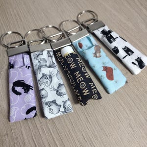 May include: Five fabric lip balm holders with key rings. The holders feature various cat-themed patterns, including black cats on white, gray cats on white, and a lavender background with black cat silhouettes. One holder has the text "MEOW MEOW".