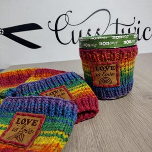 May include: Three rainbow-colored knitted cup sleeves featuring a square brown leather patch with the text LOVE is love and a rainbow icon. One sleeve is wrapped around a green nondairy container, while two others are folded in the foreground.