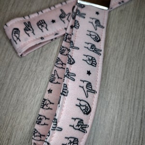 May include: Pink fabric keychain with a silver key ring. The fabric has a repeating pattern of black hand signs and stars.