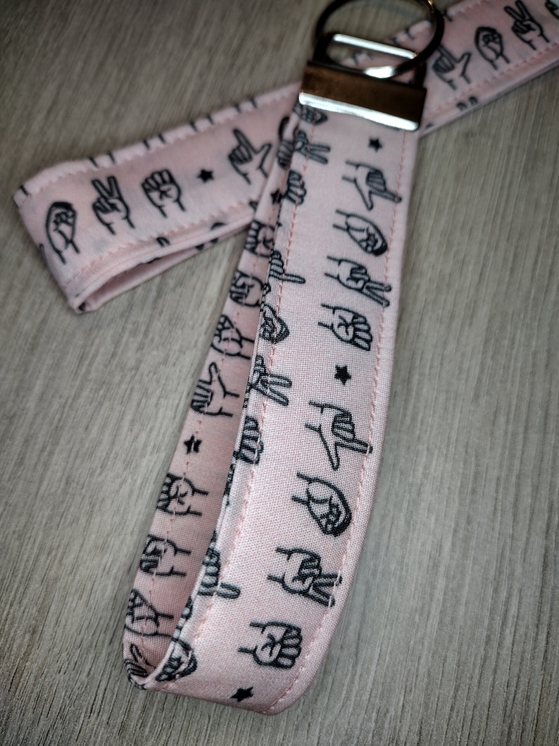 May include: Pink fabric keychain with a silver key ring. The fabric has a repeating pattern of black hand signs and stars.