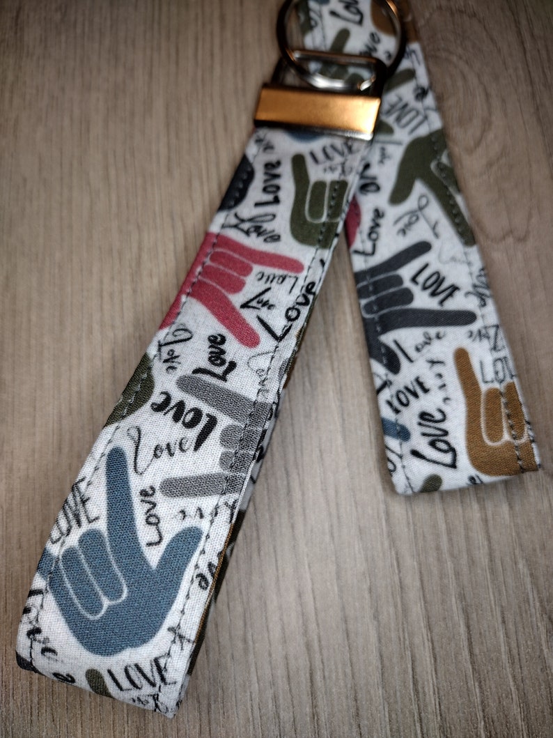 May include: A white fabric keychain with a silver key ring. The fabric is printed with a pattern of hand gestures and the word "Love" in various colors and fonts.