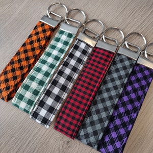 May include: Six fabric key fobs in a row, each with a different color and pattern. The patterns are all gingham, in orange and black, green and white, black and white, red and black, gray and black, and purple and black. Each has a silver metal ring.