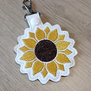 May include: Sunflower keychain with a white scalloped edge, yellow petals, and a brown center. The keychain has a silver clasp and a white leather strap with a snap closure. The design is embroidered.