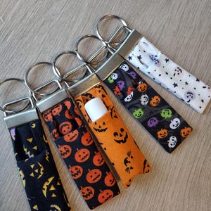 May include: Five Halloween-themed lip balm holders with key rings. The holders feature various patterns, including jack-o'-lanterns, ghosts, and stars, in black, orange, and white fabrics. One holder contains a white lip balm tube.