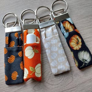 May include: Four fabric keychains with silver metal hardware. The keychains feature pumpkin patterns in orange, white, and beige on a dark gray, orange, and beige background. One keychain holds a white lip balm tube.
