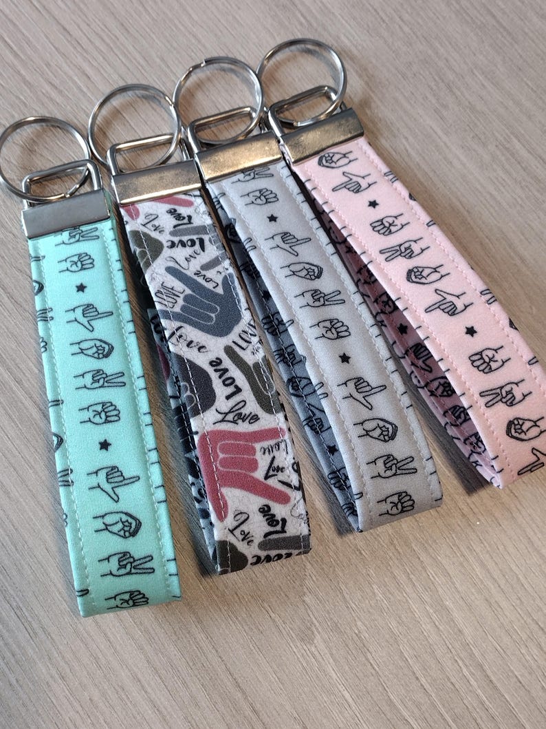 May include: Five keychains with different patterns. The keychains are made of fabric and have a silver metal ring. The patterns include hand gestures, stars, and the word "love".