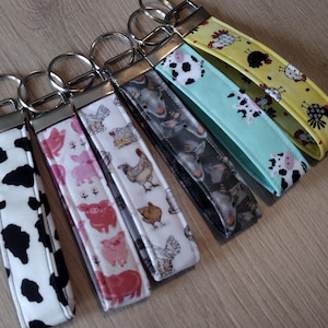 May include: A collection of fabric keychains with various farm animal patterns. Designs include black and white cow spots, pink pigs, chickens, and gray farm animal prints. Each keychain has a silver metal ring and a fabric loop.