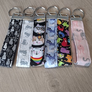 May include: Six colorful cat-themed keychains with silver key rings. The keychains are made of fabric and feature different cat designs, including a cat with sunglasses, a cat with a rainbow, and a cat with a flower crown.