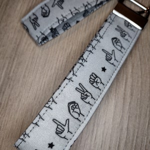 May include: A gray fabric keychain with black hand signs printed on it. The keychain has a silver metal ring and a silver metal clasp.