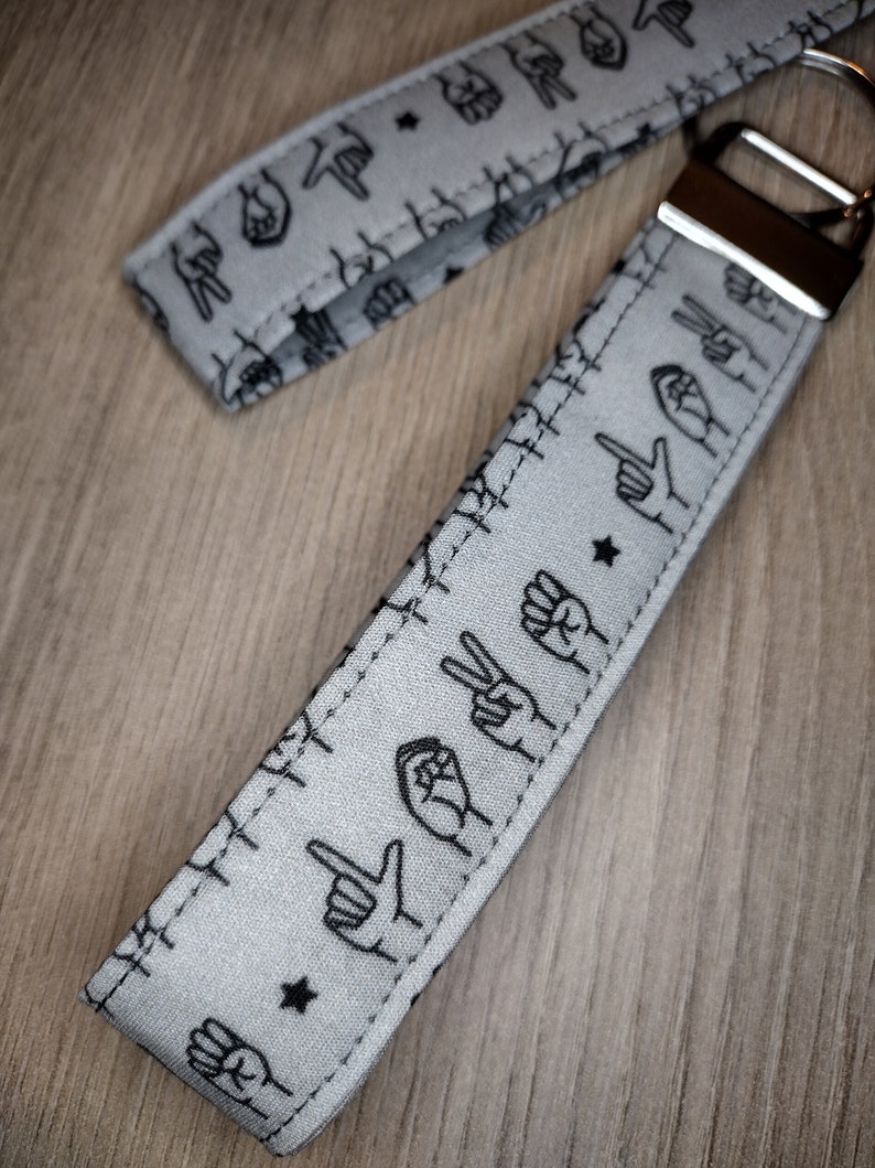 May include: A gray fabric keychain with black hand signs printed on it. The keychain has a silver metal ring and a silver metal clasp.