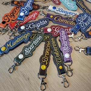 May include: A collection of embroidered keychains in various colors, featuring team names like "Tigers," "Mustangs," and "Spartans." Each keychain has a metal clasp and a button detail, perfect for sports fans.