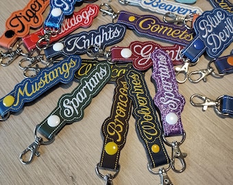 May include: A collection of embroidered fabric keychains featuring various team names such as Trojans, Bulldogs, Panthers, Colts, Bearcats, Beavers, Comets, Blue Devils, Knights, Giants, Mustangs, Spartans, Broncos, and Wolverines with metal clips.