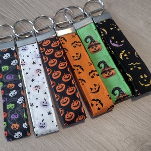 May include: Six Halloween-themed keychains with different pumpkin and witch designs. The keychains are made of fabric and have a silver metal ring.