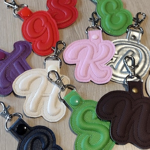 May include: A collection of letter-shaped keychains in various colors including red, green, pink, white, purple, silver, brown, and black. Each keychain features a stitched letter, a matching snap button, and a silver-toned metal lobster clasp.