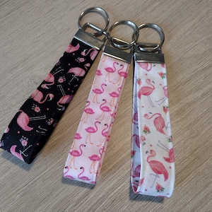May include: Three fabric keychains with metal key rings. Each keychain features a different flamingo pattern: black with pink flamingos, pink with flamingos, and white with pink flamingos and flowers.