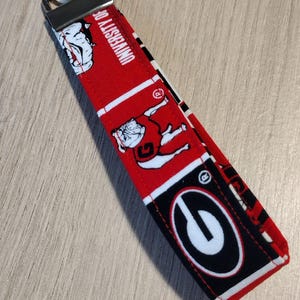 May include: A red and black fabric keychain featuring the University of Georgia logo and mascot. The keychain has a metal ring and a rectangular fabric strap with repeating patterns of the school's logo and bulldog mascot.