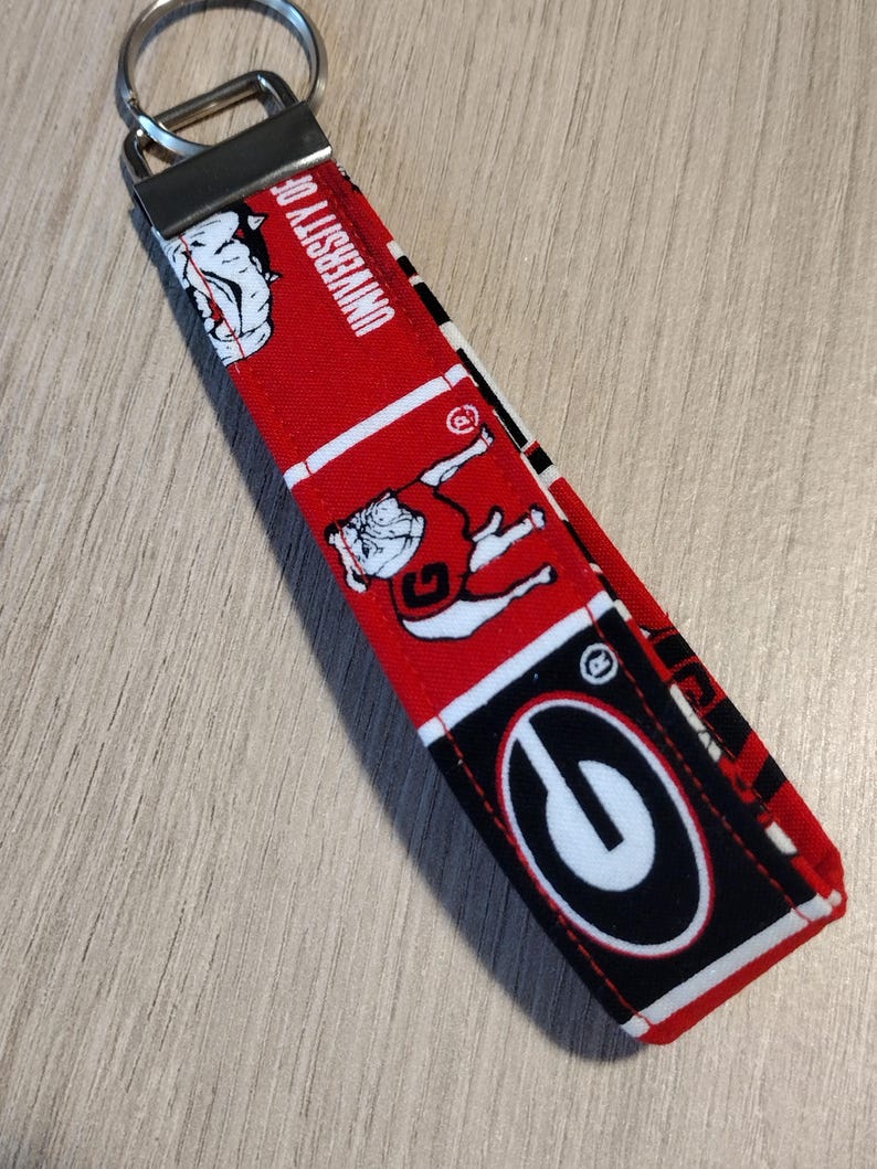 May include: A red and black fabric keychain featuring the University of Georgia logo and mascot. The keychain has a metal ring and a rectangular fabric strap with repeating patterns of the school's logo and bulldog mascot.