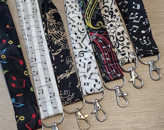 Op de afbeelding: Een collectie van zes stoffen lanyards met diverse patronen van muzieknoten en notenbalken in zwart, wit, rood, geel en groen. Elke band is afgewerkt met een zilverkleurige metalen draaibare karabijnhaak om voorwerpen veilig te bevestigen.