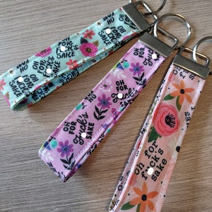 May include: Three fabric wristlet keychains with silver-tone metal hardware and split rings. The fabric features a repeating floral pattern in pink, orange, and green, interspersed with the black text "OH FOR FUCK'S SAKE" on light blue and pink backgrounds.