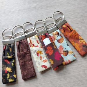 May include: A collection of six fabric lip balm holders with keychains. The holders feature various fall-themed patterns, including leaves and acorns, in colors like black, brown, orange, and cream. One holder contains a white lip balm tube.
