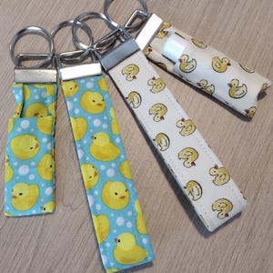 May include: Four keychains with fabric straps featuring yellow rubber duck patterns. Two keychains have a light blue background with white polka dots, while the other two have a cream background. Each keychain has a silver metal ring.