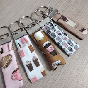 May include: Five fabric keychains with metal key rings. The keychains feature various coffee-themed patterns, including coffee cups, coffee beans, and the word "Coffee". The keychains are in shades of pink, brown, and white.