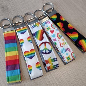 May include: Five rainbow-themed keychains with different designs. The keychains feature a rainbow stripe pattern, a rainbow heart, a peace sign, a rainbow heart with a peace sign, and a rainbow heart with the text "Love Wins".