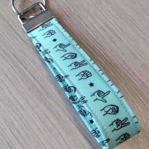 May include: A light blue fabric keychain with a silver key ring. The fabric has a repeating pattern of black hand gestures.