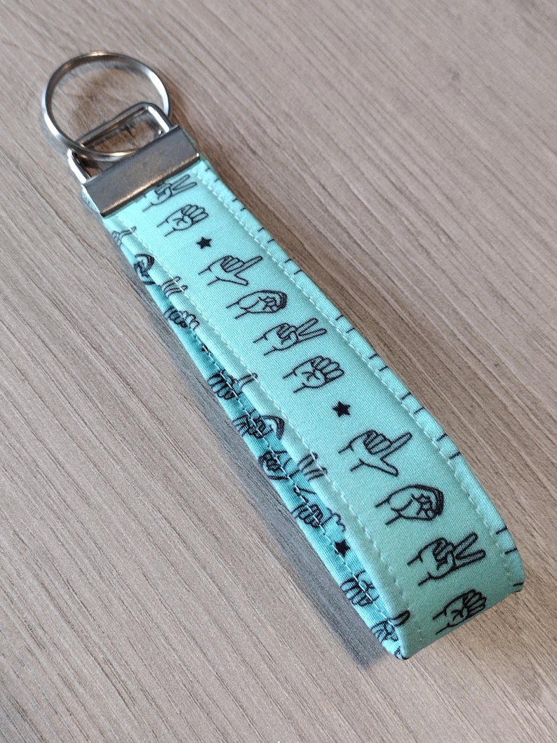 May include: A light blue fabric keychain with a silver key ring. The fabric has a repeating pattern of black hand gestures.