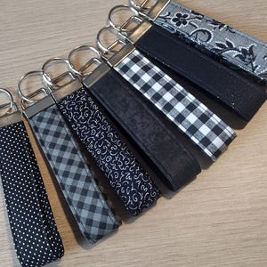 May include: A collection of seven fabric wristlet keychains featuring various black and white patterns including polka dots, gingham, floral, and solid black textures. Each keychain is attached to a silver-toned metal hardware clasp and a split ring.