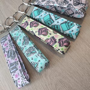 May include: A collection of fabric keychains with silver metal rings. The keychains feature book-themed patterns in various colors, including light blue, pink, and purple. Some keychains have text that reads "Books are magic" and "Just one more chapter."