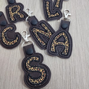 May include: A collection of personalized keychains featuring letters of the alphabet. Each keychain has a dark brown leather backing with a leopard print letter. The letters are outlined with a black stitch and attached to a silver-colored clasp.