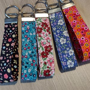 May include: Five colorful fabric keychains with floral patterns. The keychains are attached to silver key rings. The keychains are black, teal, pink, purple, and orange.
