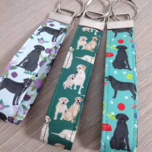 May include: Three fabric keychains with metal rings. One keychain has a light blue background with black dog silhouettes and purple flowers. The second keychain has a green background with images of golden retrievers. The third keychain has a teal background with black dog silhouettes.