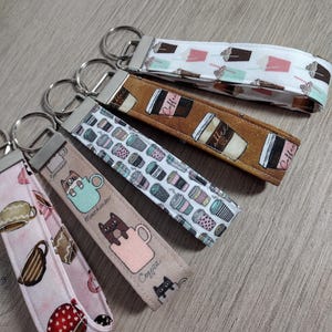 May include: A collection of fabric keychains with various coffee-themed patterns. Designs include coffee cups, coffee beans, and cats in mugs. Each keychain has a silver metal ring for attaching keys.