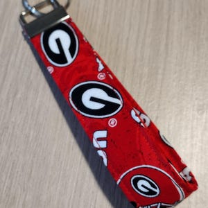 May include: Red fabric keychain with a repeating pattern of the University of Georgia Bulldogs logo and helmet. The keychain has a silver metal key ring and a black metal clasp.