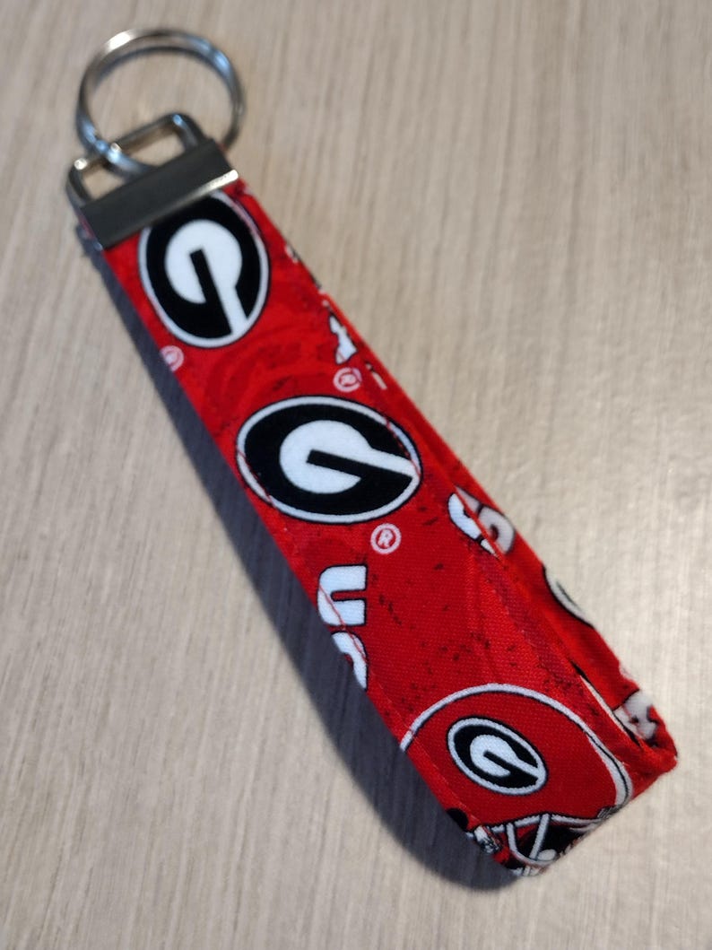 May include: Red fabric keychain with a repeating pattern of the University of Georgia Bulldogs logo and helmet. The keychain has a silver metal key ring and a black metal clasp.