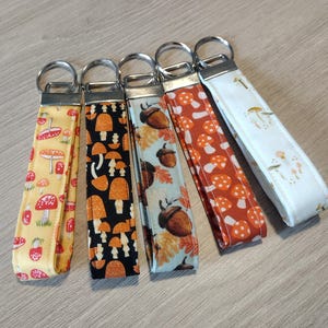May include: Five colorful keychains with different autumn-themed patterns. The keychains feature mushrooms, acorns, and leaves in shades of orange, yellow, brown, and blue.