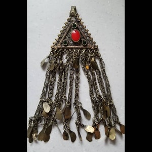 May include: A triangular pendant with a red and green stone design, featuring a chain with multiple gold-colored tassels.