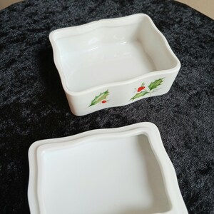 May include: A white ceramic trinket box with a wavy edge and a matching lid. The box is decorated with green holly leaves and red berries. The box is open, revealing the interior.