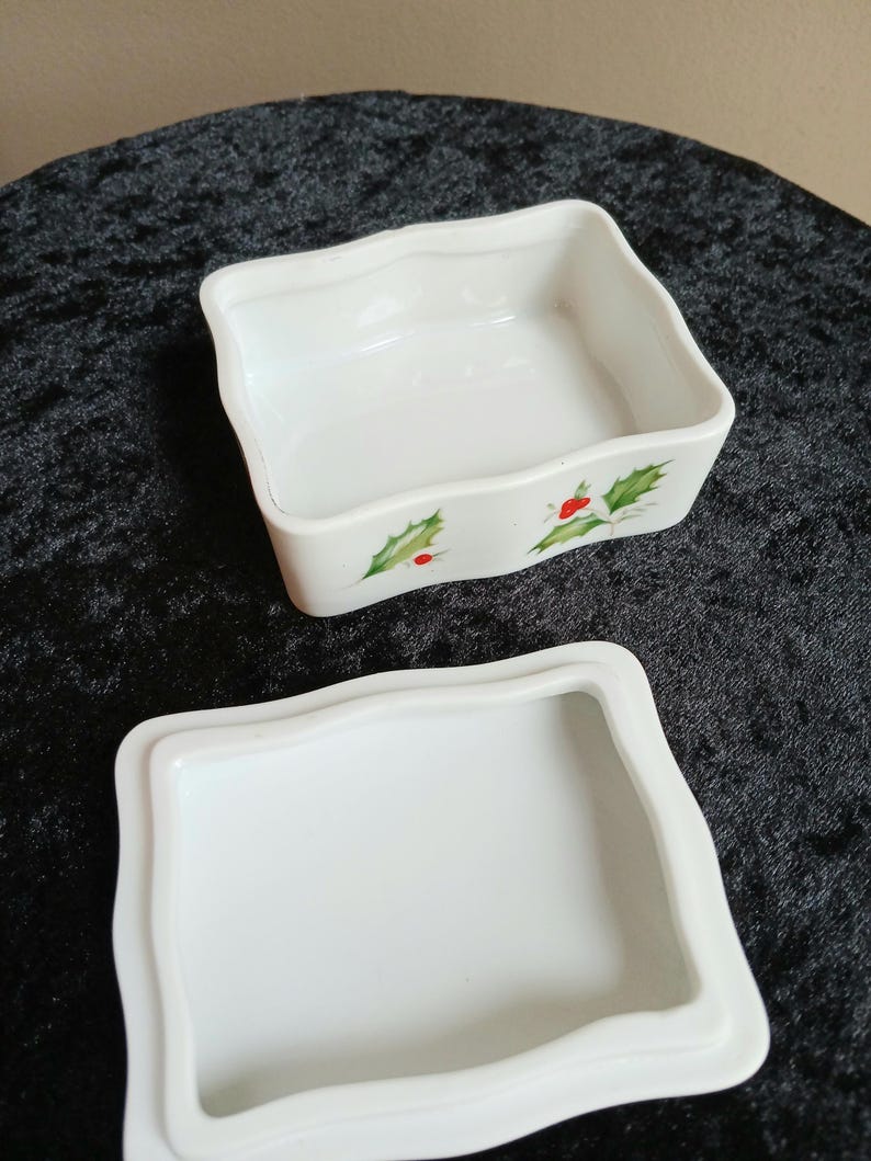 May include: A white ceramic trinket box with a wavy edge and a matching lid. The box is decorated with green holly leaves and red berries. The box is open, revealing the interior.