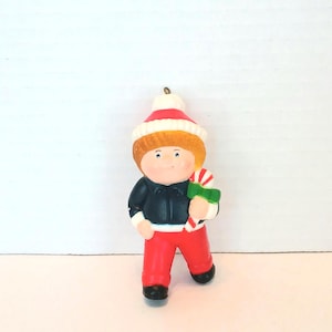 May include: A Christmas ornament of a child figure. The figure wears a red and white hat, a black jacket, and red pants. The child holds a green gift box and a candy cane. The ornament has a white background.