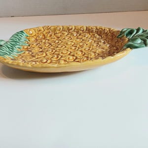 May include: A ceramic pineapple-shaped serving dish. The dish is yellow with a textured surface, mimicking the fruit's skin. Green leaves accent the ends. This decorative dish is ideal for serving snacks or as a kitchen accent.