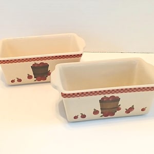 May include: Two off-white ceramic loaf pans with a red and white checkered border. Each pan features a painted design of a basket filled with red apples and scattered apples. The pans have curved handles.