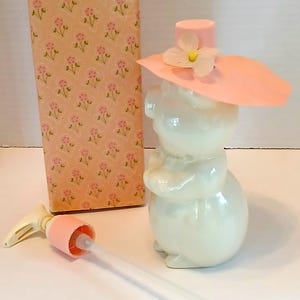 May include: A vintage perfume bottle shaped like a white bunny wearing a pink hat with a white flower. The bottle has a matching pink pump and is next to a floral-patterned box.