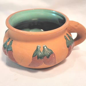 May include: A terracotta-colored ceramic bowl with a handle, featuring a green interior and red chili pepper designs with green stems. The bowl is round with a wide opening, suitable for serving soups or stews.