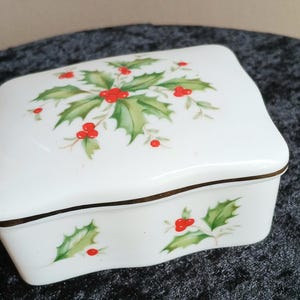 May include: A white ceramic jewelry box with a lid featuring a holly and berry design. The box has a gold trim and a curved shape. The holly leaves are green, and the berries are red. The box is likely for storing small items.