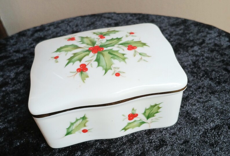 May include: A white ceramic jewelry box with a lid featuring a holly and berry design. The box has a gold trim and a curved shape. The holly leaves are green, and the berries are red. The box is likely for storing small items.