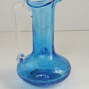 May include: A small, blue glass pitcher with a clear handle. The pitcher has a crackle finish and a flared rim. The handle is attached near the top and bottom of the pitcher. The pitcher is likely used for serving beverages.